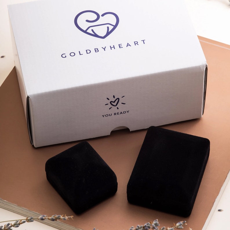 May include: A white box with the text "GOLD BY HEART" and a small heart with a sunburst design. The text "YOU READY" is below the heart. The box is sitting on a brown surface with two black velvet boxes.