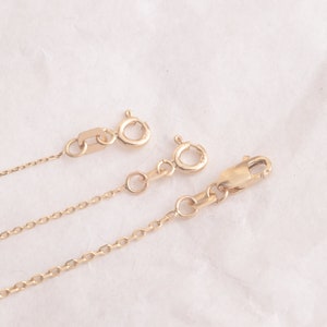 May include: Three gold chains with lobster clasps. The chains are of different lengths and are arranged in a row on a white surface.