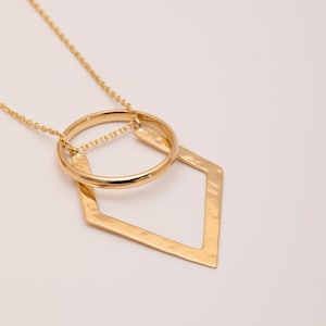 Könnte beinhalten: Eine goldene Halskette mit einem geometrischen Anhänger in Form eines Kreises und einer Raute. Der Anhänger hat eine gehämmerte Textur.