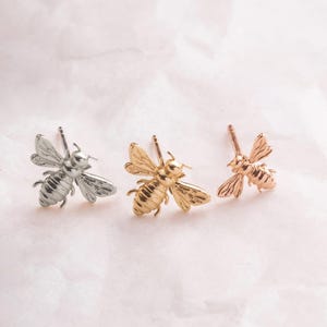 May include: Three pairs of bee stud earrings in silver, gold, and rose gold.