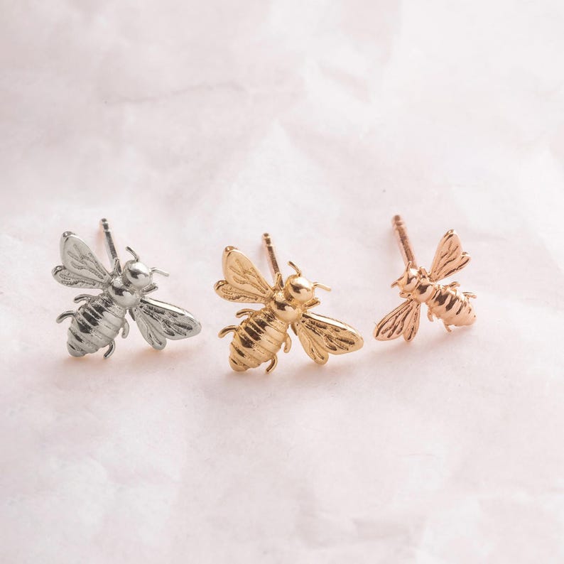 May include: Three pairs of bee stud earrings in silver, gold, and rose gold.