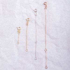 May include: Three gold chain extenders of different lengths. The extenders are made of a delicate chain with a lobster clasp on one end and a jump ring on the other.
