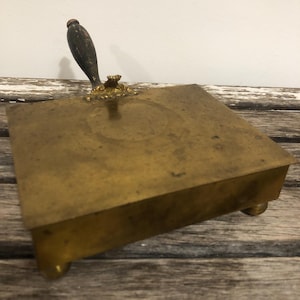 May include: A vintage brass crumb scraper with a wooden handle and decorative brass accents. The square-shaped scraper rests on small ball feet, showcasing an antique aesthetic. The handle is dark brown and the brass has a warm, aged patina.