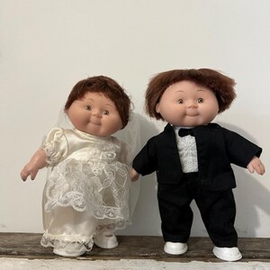 May include: Two vintage dolls dressed as a bride and groom. The bride doll wears a satin wedding dress with lace details and a veil. The groom doll is in a black tuxedo with a white shirt and bow tie. Both dolls have brown hair and rosy cheeks.
