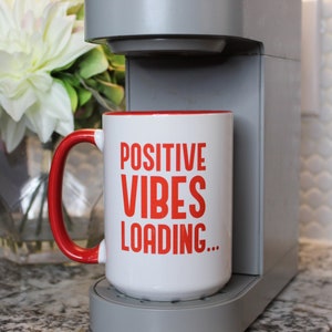 May include: A white ceramic mug with a red interior and handle. The mug features the words "POSITIVE VIBES LOADING..." in red block letters. The mug is positioned next to a gray coffee maker, with a blurred floral arrangement in the background.