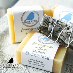May include: Three bars of handmade soap with a white label that reads "Lemongrass + Sage Sacred Smudge Soap" and "Artisan Handmade Cold-Process | Vegan All Natural Soap Bar". The soap is a light yellow color and is sitting on a white surface with a bundle of sage tied with white string.