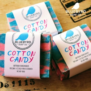 May include: Three bars of soap with a blue and pink swirl design. Each bar has a white label with the text "BLUEBYRD SOAP CO." and "COTTON CANDY". The label also includes the text "ARTISAN HANDMADE", "VEGAN", "COLD-PROCESSED", and "SOAP BAR".