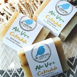 May include: Three bars of handmade soap with a bluebird logo. The soap is labeled "Aloe Vera Calendula" and is described as artisan handmade, cold-process, and vegan.