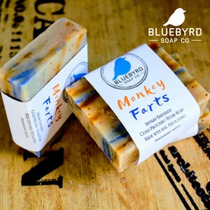May include: Two bars of handmade soap with a blue and yellow swirl design. The soap is labeled "Monkey Farts" and is made by Bluebyrd Soap Co. The label also states that the soap is artisan handmade, cold-process, vegan, and made with real exfoliating strawberry seeds.