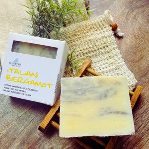 May include: A bar of handmade, vegan Italian Bergamot soap. The soap is white with gray swirls and is sitting on a wooden soap dish. The soap is packaged in a white box with the brand name "Bluebyrd Soap Co." and the text "Italian Bergamot" in yellow.