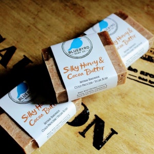 May include: Three bars of handmade soap with a bluebird logo and the text "Silky Honey & Cocoa Butter" on the label. The soap is made with organic cocoa butter, oatmeal, cinnamon, and honey.