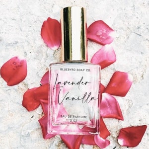 May include: A clear glass bottle of lavender vanilla perfume with a gold cap and label. The bottle is sitting on a white surface with pink rose petals scattered around it. The label reads "Bluebyrd Soap Co. Lavender Vanilla Eau de Parfum 2 oz."