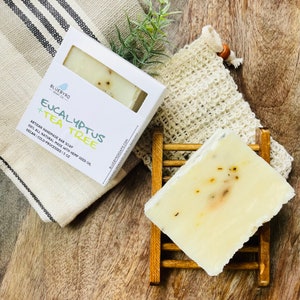 May include: A bar of handmade soap with a white base and flecks of green and yellow. The soap is labeled "Eucalyptus + Tea Tree" and is made with hemp seed oil. The soap is on a wooden soap dish with a white washcloth and a mesh soap bag.
