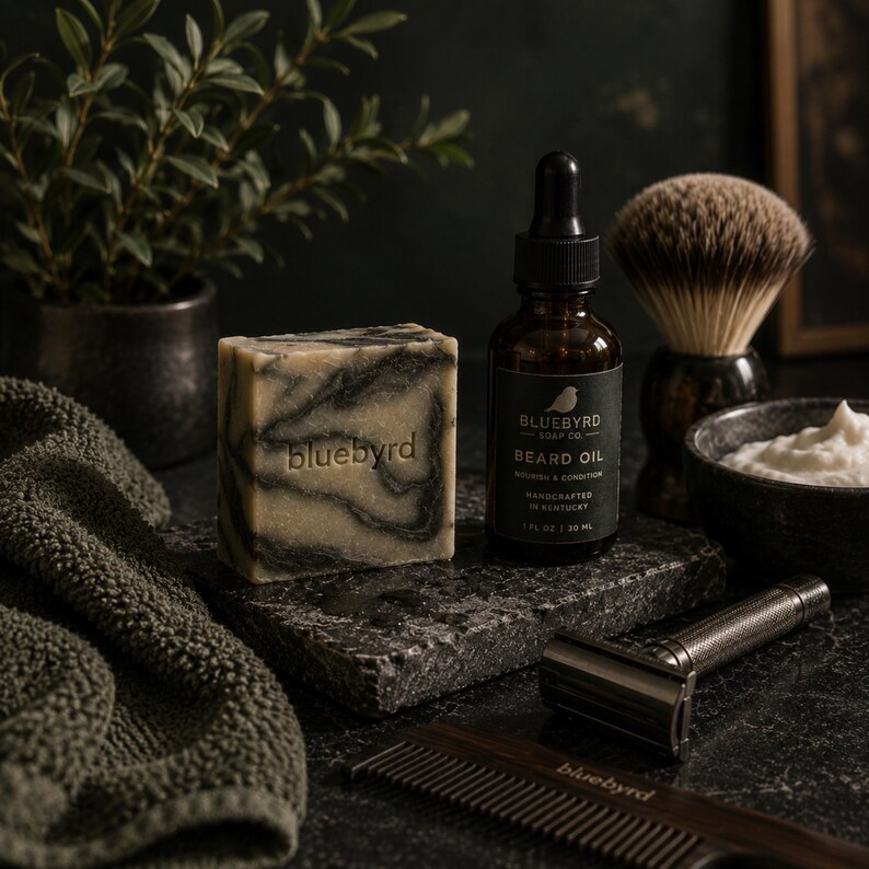May include: A bar of marbled black and beige soap with the word bluebyrd embossed, a brown glass bottle of beard oil labeled Bluebyrd Soap Co. 1 fl oz 30 ml, a shaving brush, a metal safety razor, a wooden comb, and a dark green textured towel.