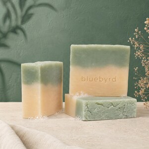 May include: Three rectangular bars of soap with a dual-tone design featuring a sage green top and a cream-colored bottom. The largest bar is embossed with the text bluebyrd in lowercase letters. Small soap bubbles are visible at the base.