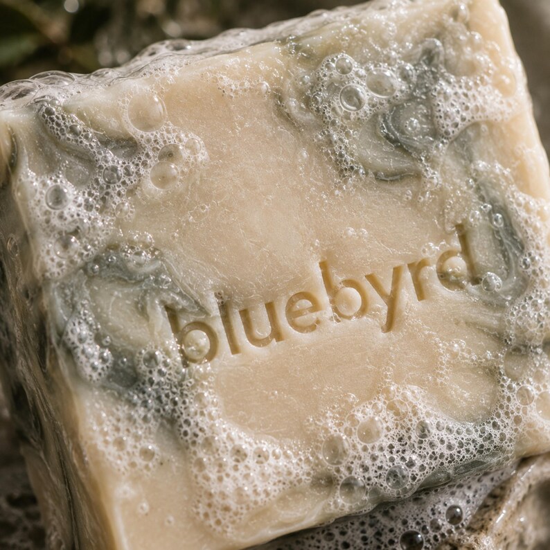 May include: A square bar of beige soap with a marbled grey pattern and the word bluebyrd embossed in the center. The surface of the soap is covered in a layer of white bubbly lather, highlighting the texture and the stamped lettering on the bar.