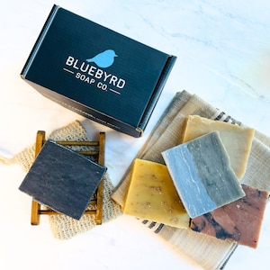 May include: A black box with the text "Bluebyrd Soap Co." and a blue bird logo. Four bars of soap are arranged on a beige towel. The soap bars are in shades of gray, brown, and yellow.