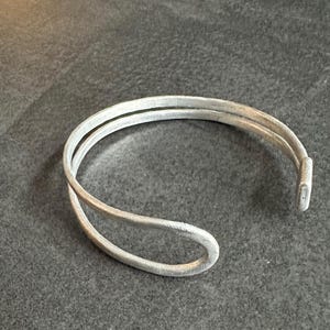Può includere: Un bracciale a polsino color argento con un design a doppia fascia. Il bracciale ha un design semplice e aperto con una leggera curva ed è realizzato in materiale metallico. Il bracciale è mostrato su una superficie grigio scuro.