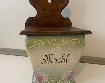 Puede incluir: Especiero de pared de madera antigua con un recipiente de cerámica. El recipiente está decorado con un diseño verde y blanco y la palabra "Mehl" en escritura negra. El tablero trasero de madera tiene tallas decorativas.