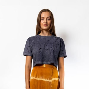 May include: A woman is wearing a dark gray cropped t-shirt with a tie-dye pattern and a long, orange and white tie-dye skirt. She is wearing light yellow sandals.