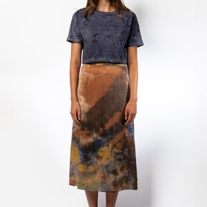 May include: A woman is wearing a dark gray cropped t-shirt with a tie-dye pattern and a brown, orange, and blue tie-dye midi skirt. She is wearing yellow sandals.