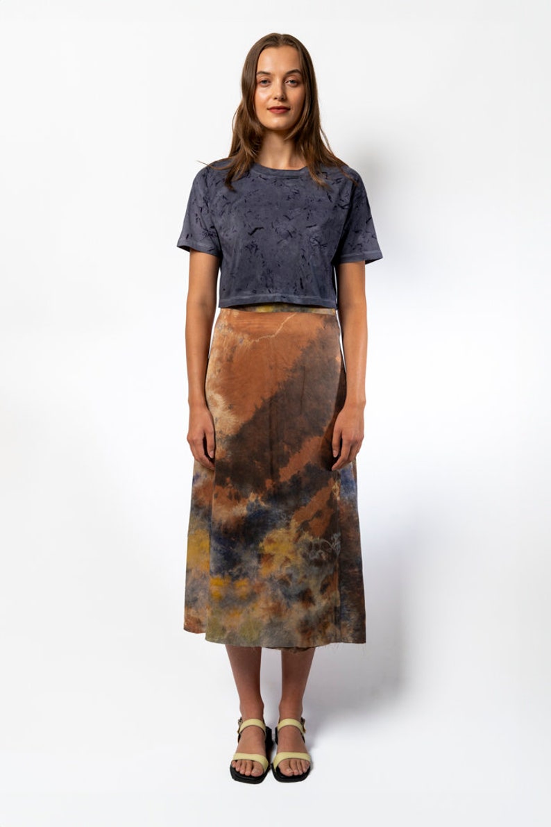 May include: A woman is wearing a dark gray cropped t-shirt with a tie-dye pattern and a brown, orange, and blue tie-dye midi skirt. She is wearing yellow sandals.
