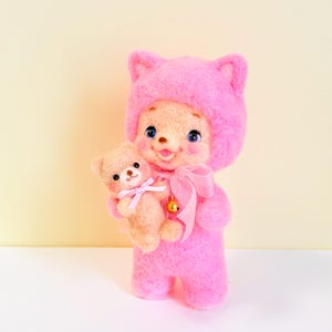 以下が含まれることがあります： 猫耳付きの鮮やかなピンク色のフード付きロンパースを着たニードルフェルト人形。青い目と開いた口が特徴です。人形は、首にピンクのリボンと金の鈴をつけた、小さな薄茶色のニードルフェルトのテディベアを抱えています。