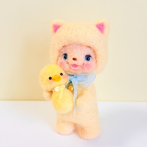 May include: A needle-felted cream-coloured cat figurine wearing a hood with pink inner ears, blue eyes, and rosy cheeks. The cat holds a small yellow chick with a tiny yellow ribbon around its neck. A light blue sheer ribbon with a small gold bell is tied around the cat's neck.