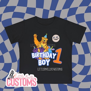 May include: Black t-shirt with a colorful graphic design for a first birthday. The design includes a waving bear, other cartoon characters, the words "BIRTHDAY BOY" and the number "1". The shirt also has the text "LITTLEBELLECUSTOMS".