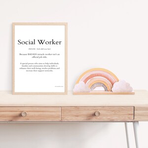 May include: A framed print with a white background and black text that reads "Social Worker" and a definition of the word. The print is on a wooden desk with a rainbow-coloured wooden toy and two white clouds.
