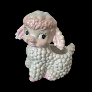 May include: A ceramic lamb figurine with a textured, cream-colored wool body and a smooth, pale pink head. The lamb features large, dark eyes with long eyelashes, a small smile, and pink-tinted ears. An open cavity is located on the back.