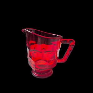 May include: A translucent ruby red glass pitcher featuring a faceted geometric exterior pattern, a wide pouring spout, a sturdy handle, and a circular pedestal base. The vessel is crafted from pressed glass with a vibrant, deep red color throughout.