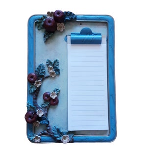 May include: A decorative clipboard with a blue frame and a light blue background. The clipboard features a branch with red apples and white flowers. A pad of lined paper is held in place by a blue clip.