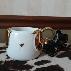 May include: White ceramic mug shaped like a dog with gold ears and nose. The mug has a gold handle and is attached to a black leather dog keychain with a gold tassel.