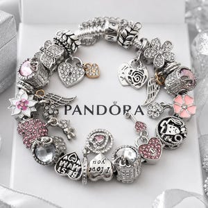 Può includere: Un bracciale con ciondoli in argento con vari elementi tra cui cuori, fiori, ali e un orsetto, con cristalli rosa e trasparenti. Diversi ciondoli riportano incisioni come I love you e Best Mum. Il logo Pandora è chiaramente visibile.