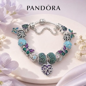 Può includere: Un bracciale Pandora in argento con vari charm colorati, tra cui farfalle viola e verdi, sfere testurizzate color ottanio e un ciondolo centrale a forma di cuore con una pietra viola e un dettaglio a fiocco in argento sovrapposto.