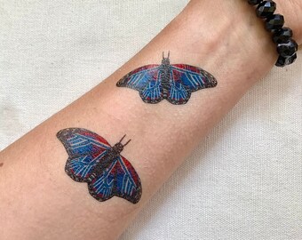 May include: Two temporary butterfly tattoos on a person's arm. The butterflies are blue, red, and black with intricate patterns.