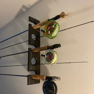 May include: A dark brown wooden fishing rod rack mounted on a white wall. The rack holds three fishing rods with reels. The reels are green, black, and blue. The rods are thin and black.