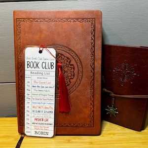 May include: A white bookmark with a red tassel, labeled "Book Club Reading List" with a list of book titles for each month of the year. The bookmark is attached to a brown leather journal with a decorative design.