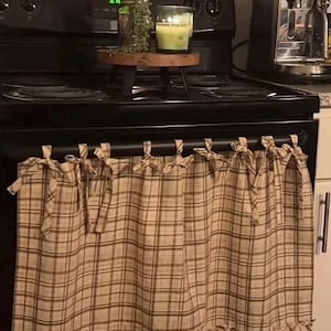 May include: A beige and brown plaid fabric curtain with a ruffled bottom edge, attached to a black oven handle with multiple tied fabric loops. The curtain features a grid pattern of intersecting lines and hangs straight across the front.