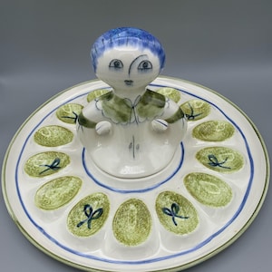 May include: A white ceramic egg holder with a green and blue design. The holder has 12 individual egg-shaped compartments and a removable lid featuring a stylized figure with blue hair and a green dress.