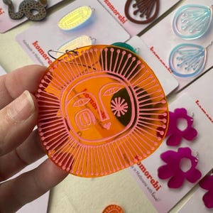 May include: A collection of colorful earrings, including a large orange sun-shaped earring with pink detailing. Other earrings include flower, snake, and abstract designs. The earrings are displayed on white cards with the word "komorebi".