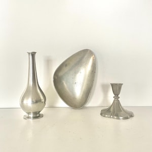 Puede incluir: Colección de tres objetos decorativos de metal plateado. Incluye un jarrón delgado, un plato de forma única y un pequeño candelabro. Los objetos están dispuestos sobre una superficie blanca.