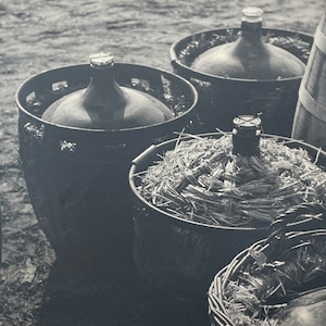 May include: A black and white image showing several large glass bottles, some in metal baskets and others in woven containers. The bottles have rounded bodies and are topped with stoppers. Some baskets are filled with straw.