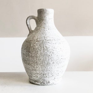 以下が含まれることがあります： テクスチャ加工された表面とハンドルが付いた白いセラミックの花瓶。花瓶は丸みを帯びたボディと狭い首を持っています。