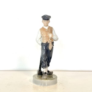 May include: A porcelain figurine of a man in traditional attire. He wears a dark cap, a tan waistcoat over a white shirt, and dark blue trousers. He holds a tool and a bag, standing on a round base. The figurine is a collectible item.