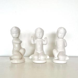 Könnte beinhalten: Drei weiße Keramikfiguren von Babys in verschiedenen Posen. Eine hat verschränkte Arme, eine andere hat erhobene Arme und die dritte kniet. Jede Figur steht auf einem runden Sockel und hat eine glänzende Oberfläche.