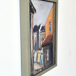 May include: A framed oil painting depicting a street scene with colorful buildings and laundry hanging on a line. The painting is in a light green frame.
