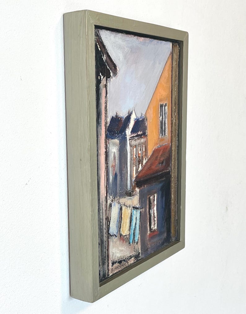 May include: A framed oil painting depicting a street scene with colorful buildings and laundry hanging on a line. The painting is in a light green frame.