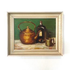 May include: Oil painting depicting a still life with a copper tea kettle, a dark bottle, a small mug, and a bottle stopper. The objects are set against a green backdrop and rest on a white surface. The painting is framed in a gold-coloured frame.